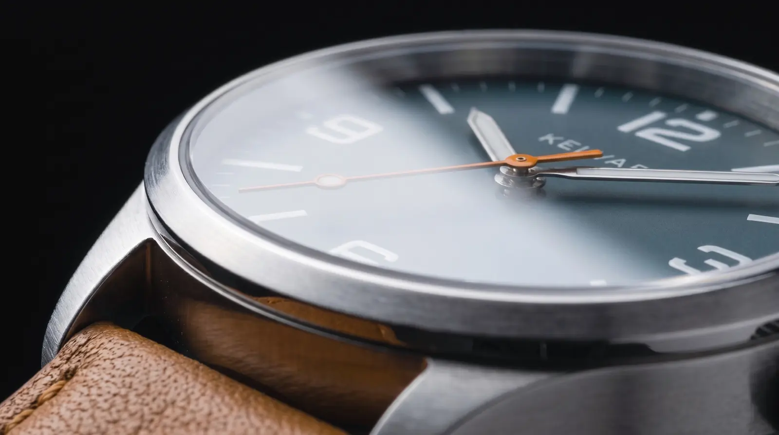 Low view across the crystal: a band of light pales the dial, the bevel holds a bright edge and the orange seconds hand runs toward the 9.