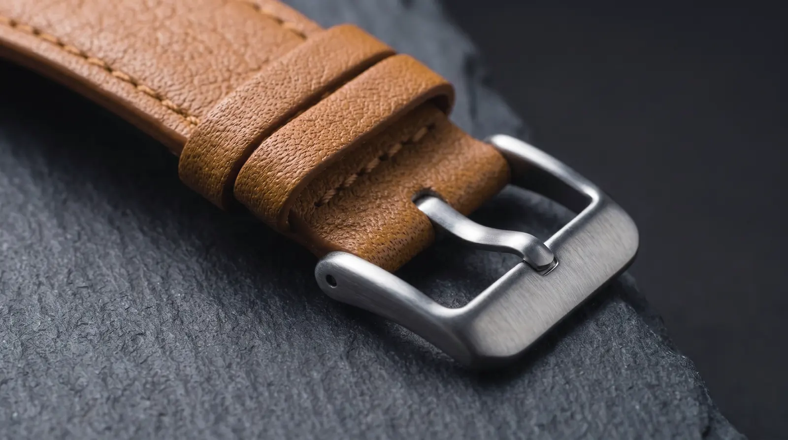 The tan grained strap with tonal stitching, two keepers and a pin buckle, resting on slate.